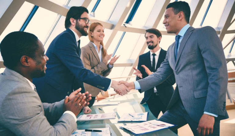 Un gruppo di persone sedute attorno a un tavolo in un ambiente formale. Due di loro si stringono la mano dopo aver firmato un contratto, mentre gli altri osservano.